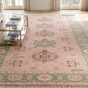 May include: A large rectangular area rug featuring a soft pink central field with repeating geometric motifs in sage green and cream. The rug has a wide sage green border with floral patterns and a fringed edge. The text pbrugscreations is visible.