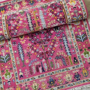 May include: A rolled-up, rectangular, pink rug with a detailed floral and tree design. The rug features a border with repeating patterns in shades of blue, yellow, green, and pink. The design includes a central tree motif and other floral elements.