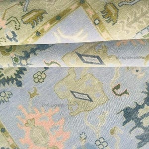 May include: Close-up of a rolled-up area rug with a detailed, traditional design. The rug features a light blue background with intricate patterns in shades of green, peach, and navy. The design includes floral and geometric elements.