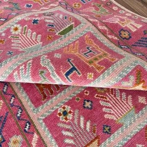 Op de afbeelding: Een roze tapijt met een geometrisch patroon met gestileerde dieren en planten. Het tapijt heeft een lichtblauwe en witte rand.