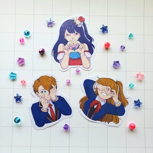 Może przedstawiać: Trzy naklejki w stylu anime przedstawiające postacie w różnych pozach. Jedna naklejka przedstawia postać tworzącą serce dłońmi. Dwie pozostałe naklejki przedstawiają postacie z gestami dłoni w pobliżu oczu. Naklejki otoczone są kolorowymi koralikami w kształcie gwiazd.
