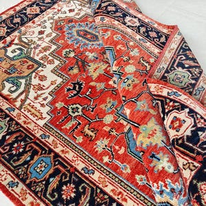 Puede incluir: Una alfombra enrollada con un diseño intrincado, dominada por un campo central rojo. La alfombra presenta detalles en crema, azul marino y azul. El borde es azul marino con motivos florales, y la alfombra está parcialmente desenrollada, mostrando su patrón detallado.