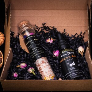 May include: A brown cardboard box filled with black shredded paper. Inside the box are two glass bottles with labels that read "Arcane Wictery" and "Mountain's Sabbath". One bottle contains bath salts and the other contains body oil.
