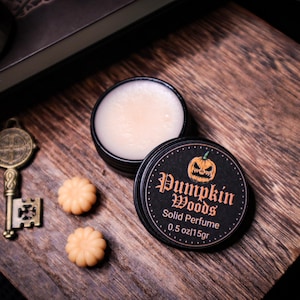 Peut inclure: Un petit pot noir de parfum solide Pumpkin Woods avec un motif de citrouille, à côté de deux fondants parfumés en forme de citrouille et d'une clé décorative. Le pot de parfum est ouvert, révélant le parfum solide.