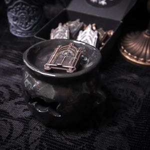 May include: A black ceramic cauldron with a gold-toned metal archway design on top. The cauldron is sitting on a black lace fabric.