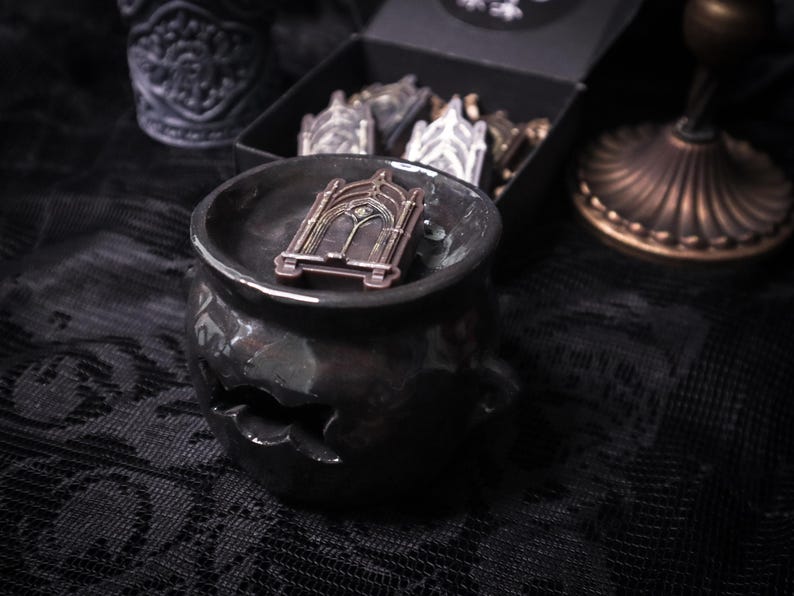 May include: A black ceramic cauldron with a gold-toned metal archway design on top. The cauldron is sitting on a black lace fabric.