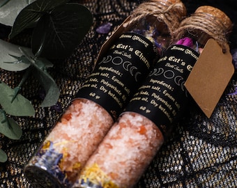 May include: Two glass bottles filled with pink bath salts, dried flowers, and crystals. The bottles have black labels with gold lettering that reads "Arcane Wiccery Black Cat's Charm Bath Salts" and "Arcane Wiccery Black Cat's Charm Bath Salts". The bottles are tied with twine and have brown paper tags.