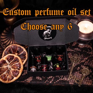 Può includere: Un set di oli profumati personalizzati in una scatola nera con la scritta "Choose any 6". La scatola contiene diverse piccole bottiglie di profumo. Sono presenti anche fette d'arancia essiccate e chiavi antiche, creando un'estetica gotica.