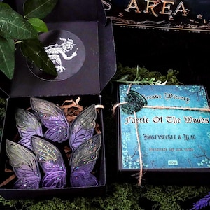 May include: A black box with six purple and green fairy wings with glitter. Beside it is a box of "Faerie Of The Woods" scented wax melts, tied with twine and sealed with a green wax seal.