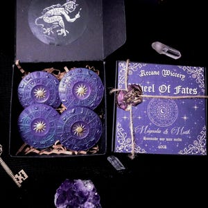 May include: A dark image showing a black box containing four purple wax melts with a sunburst design. A matching box with the text "Arcane Wiccery Wheel Of Fates" is tied with twine. Crystals and a key are also visible.