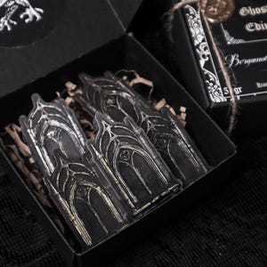 May include: A black box with a silver sticker of gothic arches. The box is open and the sticker is visible. The box is labelled "Ghosts of Edinburgh Bergamot & Oak Handmade Soy Wax 5 gr"