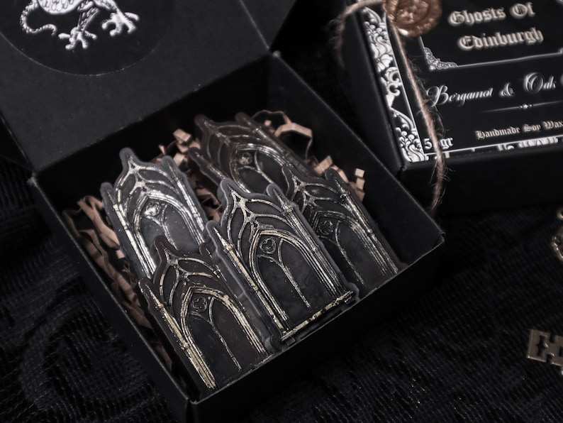 May include: A black box with a silver sticker of gothic arches. The box is open and the sticker is visible. The box is labelled "Ghosts of Edinburgh Bergamot & Oak Handmade Soy Wax 5 gr"
