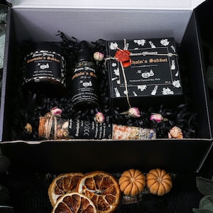 Puede incluir: Una caja de regalo negra llena de productos de baño y cuerpo con temática de Halloween. La caja contiene una vela, una botella de aceite corporal, una bolsa de sales de baño y una caja de ceras de soja hechas a mano. Los productos están todos etiquetados con las palabras "Samhain's Sabbat".