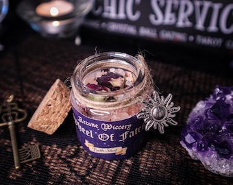 May include: A small glass jar filled with pink and white crystals and dried rose petals, labeled "Arcane Wiccery Wheel Of Fate Bath Shot." The jar is adorned with twine and a silver sun charm. A key and amethyst cluster are nearby.