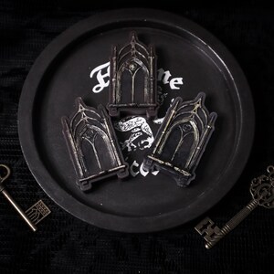 May include: Three black and silver decorative gothic archway-shaped keychains on a black tray with the word "Home" written on it.