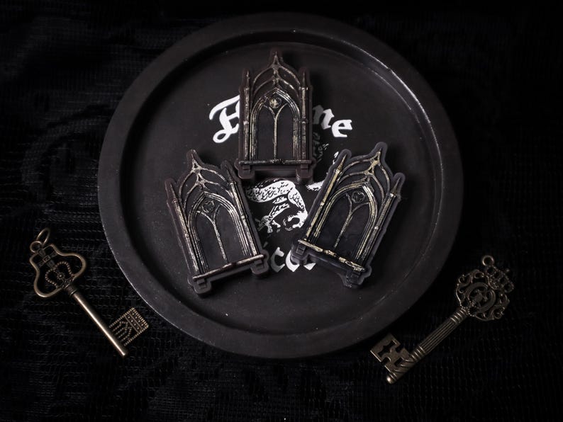 May include: Three black and silver decorative gothic archway-shaped keychains on a black tray with the word "Home" written on it.