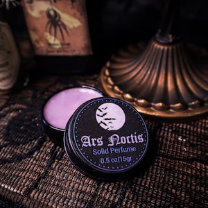 Puede incluir: Una lata negra de perfume sólido con el texto "Ars Noctis" y un diseño de luna y murciélagos. El perfume es de color morado claro y la lata contiene 15g. El fondo incluye un vaso decorativo y botellas.