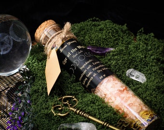 May include: A glass bottle filled with pink bath salts and labeled "Eye of the Wild" with a moon and star design. The bottle is sitting on a bed of green moss with a crystal and a key.