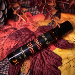 Könnte beinhalten: Eine kleine Parfümflasche mit schwarzem Deckel, beschriftet mit "Arcane Hollow Wood Perfume 10ml". Die Flasche liegt auf herbstlichen Blättern in Rot- und Gelbtönen. Das Etikett hat eine orangefarbene gotische Schrift.