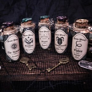 May include: Five glass potion bottles with cork stoppers and labels reading "Arcane Wiccery." Each bottle features a different label design and ingredient name, including "The Alchemy Rosemary" and "Ophis Dandelion Root." Decorative keys and crystals are also present.