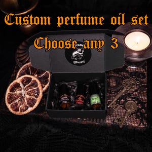 Può includere: Un set di oli profumati personalizzati in una scatola nera, con tre piccole bottiglie all'interno. La scatola è aperta, rivelando le bottiglie e l'imbottitura di carta nera. Il testo "Custom perfume oil set" e "Choose any 3" sono visualizzati. Sono presenti anche chiavi decorative e fette d'arancia essiccate.