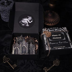 May include: Black box with a wax seal and a label that reads "Arcane Wiccery Ghosts of Edinburgh Bergamot & Oak Moss Handmade Soy Wax Melts". The box is open and contains a set of black and gold gothic arch stickers.