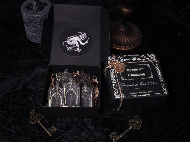 May include: Black box with a wax seal and a label that reads "Arcane Wiccery Ghosts of Edinburgh Bergamot & Oak Moss Handmade Soy Wax Melts". The box is open and contains a set of black and gold gothic arch stickers.