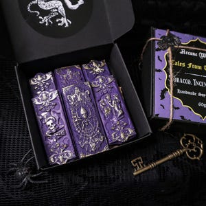 Peut inclure: Un ensemble de cinq galets de cire de soja violets et dorés dans une boîte noire. Les galets de cire sont en forme de petits livres avec des motifs complexes et le texte "Arcane Wiccery Tales From The Crypt Tobacco. Incense & Musk Handmade Soy Wax Melts 60gr" sur la boîte.