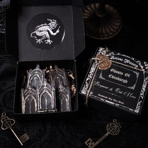 May include: Black box with a white sticker of a demon and a white sticker of a gothic archway. The box is open and contains a set of gothic archway stickers. The box is sitting on a black surface.  The text on the box reads "Arcane Wiccery Ghosts of Edinburgh Bergamot & Oak Moss Handmade Soy Wax Melts"