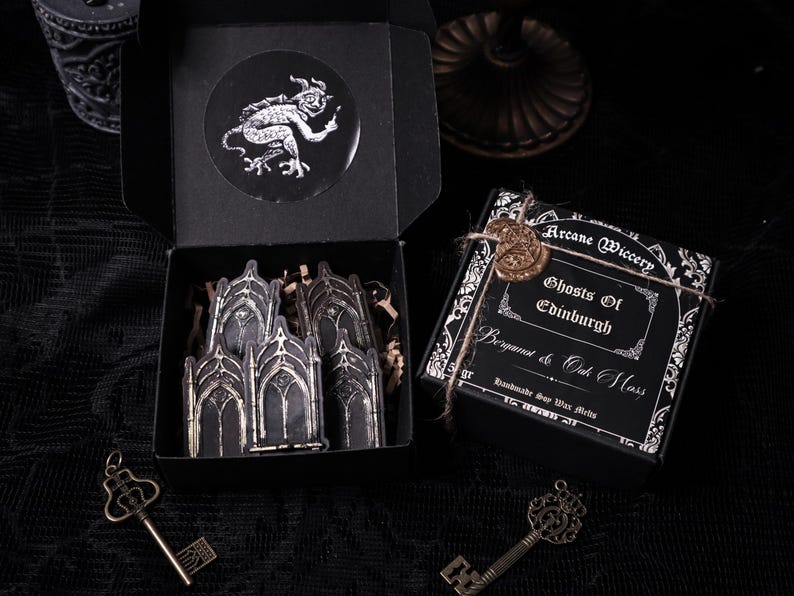 May include: Black box with a white sticker of a demon and a white sticker of a gothic archway. The box is open and contains a set of gothic archway stickers. The box is sitting on a black surface.  The text on the box reads "Arcane Wiccery Ghosts of Edinburgh Bergamot & Oak Moss Handmade Soy Wax Melts"