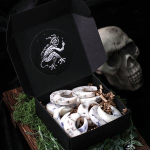 May include: A black box with a white sticker of a demon holding a lit match. The box is open and filled with white crescent moon shaped candles with dried lavender inside. The candles are sitting on brown paper shreds.