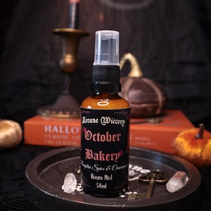 Op de afbeelding: Een 50ml amberkleurige glazen sprayfles met Arcane Wiccery October Bakery roomspray en een zwarte pompdop. Het zwarte etiket bevat rode tekst met de woorden October Bakery, Pumpkin Spice & Cinnamon, Room Mist en kleine rode esdoornbladeren.