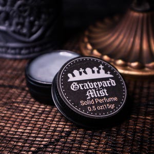 Op de afbeelding: Een zwarte blikken doos met vaste parfum, met het opschrift "Graveyard Mist" en een kerkhofontwerp. De doos is open en toont het parfum. Het etiket luidt "0,5 oz/15g". De achtergrond is donker en gestructureerd.