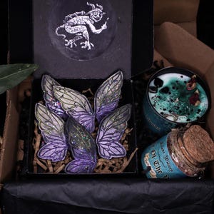 Könnte beinhalten: Eine Geschenkbox mit lila und grünen Feenflügel-Ornamenten, einer türkisfarbenen Kerze mit dekorativen Elementen und einem Glas mit Korkdeckel. Eine schwarze Box mit einer Drachenillustration ist ebenfalls sichtbar.
