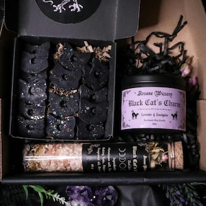 Könnte beinhalten: Eine schwarze Geschenkbox gefüllt mit schwarzen, katzenförmigen Wachs-Schmelzen, einer schwarzen Kerze mit dem Text "Arcane Wiccery Black Cat's Charm Lavender & Eucalyptus" und einem kleinen Glas mit getrockneten Kräutern und dem Text "Black Cat's Charm Rose Quartz"