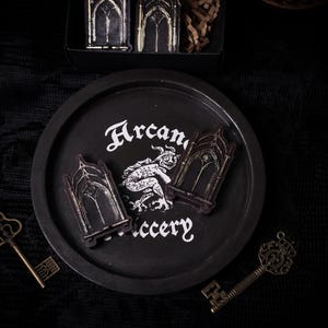 May include: Black and white illustration of a gothic archway with a small, black and white illustration of a creature inside. The illustration is on a black plate with the words "Arcanery" written in white.