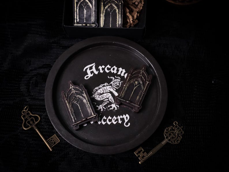 May include: Black and white illustration of a gothic archway with a small, black and white illustration of a creature inside. The illustration is on a black plate with the words "Arcanery" written in white.