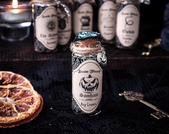 May include: A small glass jar with a cork stopper and twine, labeled "Arcane Wiccery Samhain Bay Leaves." The jar has a black and white patterned design and a pumpkin graphic. Other jars with labels are in the background, along with a lit candle and a decorative key.