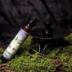 Może przedstawiać: Butelka olejku perfumowanego z etykietą ważki, zatytułowana "Night Silk", spoczywa na zielonym mchu. Obecny jest również czarny świecznik i dekoracyjne klucze. Butelka ma 10 ml. Ogólnym motywem jest mistycyzm i natura.