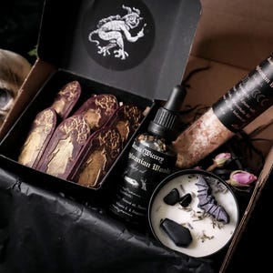 May include: A black box filled with gothic-themed items. Includes a bottle of body oil, a candle with a bat and coffin, a tube of bath salts, and coffin-shaped soaps. A skull is visible in the background. The box has a sticker with a demon.