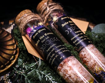 May include: Two glass test tubes filled with pink Himalayan bath salts and dried flowers. The tubes are labeled "Into the Coven" and "Into the Moon" with black and gold lettering. The labels list ingredients including Himalayan salt, Epsom salt, baking soda, polysorbate, fragrance oil, and dry botanicals. The tubes are sitting on a bed of green moss.