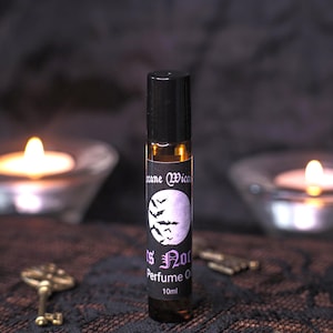 May include: A perfume oil bottle with a black cap and a label showing a purple moon and bats. The label reads "Is Not" and "Perfume Oil 10ml". The bottle is displayed against a dark backdrop with lit candles, creating a gothic aesthetic.