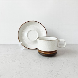 May include: A white ceramic coffee cup and saucer with a brown rim. The cup and saucer have a speckled texture.
