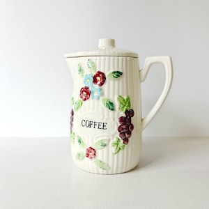 May include: Cream-colored ceramic coffee pot with a lid and handle. The pot is decorated with floral and grape designs in red, blue, and green. The word "COFFEE" is printed on the front.