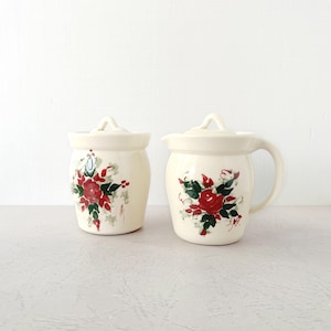 May include: Two off-white ceramic containers with lids, one with a handle. Each is decorated with a red rose and green leaf floral design. The containers are likely for kitchen use.