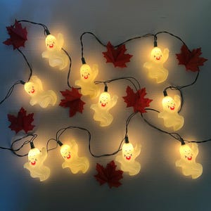 May include: A decorative string of Halloween lights. The lights feature smiling ghost-shaped bulbs, strung on black wire, and interspersed with red maple leaf decorations. The bulbs emit a warm glow, ideal for festive decorations.