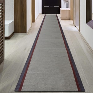 Puede incluir: Una alfombra de pasillo gris con rayas rojas y azul marino a lo largo de los bordes. La alfombra está colocada en un pasillo con suelo de madera.