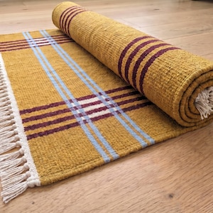 May include: A mustard yellow woven rug featuring a plaid pattern with dark maroon and light blue stripes. The rug has a textured surface and is partially rolled up, revealing white fringe tassels along the edge of the rectangular fabric.