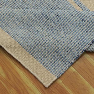 Puede incluir: Una alfombra de rayas azules y blancas con un borde crema. La alfombra está doblada por la mitad y está sobre un suelo de madera.