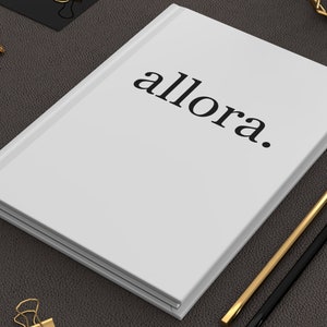 May include: A white notebook with the word "allora." printed in black on the cover. Two pencils, one gold and one black, are lying on a dark brown surface next to the notebook.